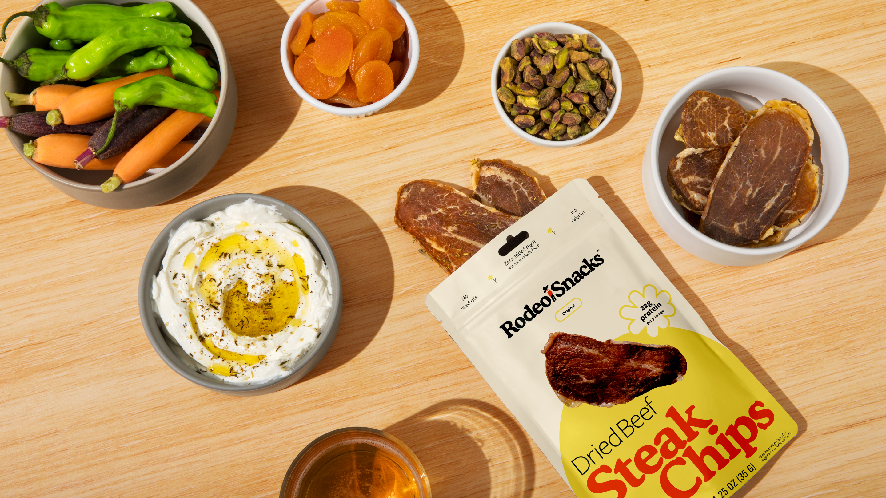 Bowls of ingredients - peppers, dried apricots, pistachios, and yogurt - on a wooden cutting board with a bag of Rodeo Snacks Steak Chips